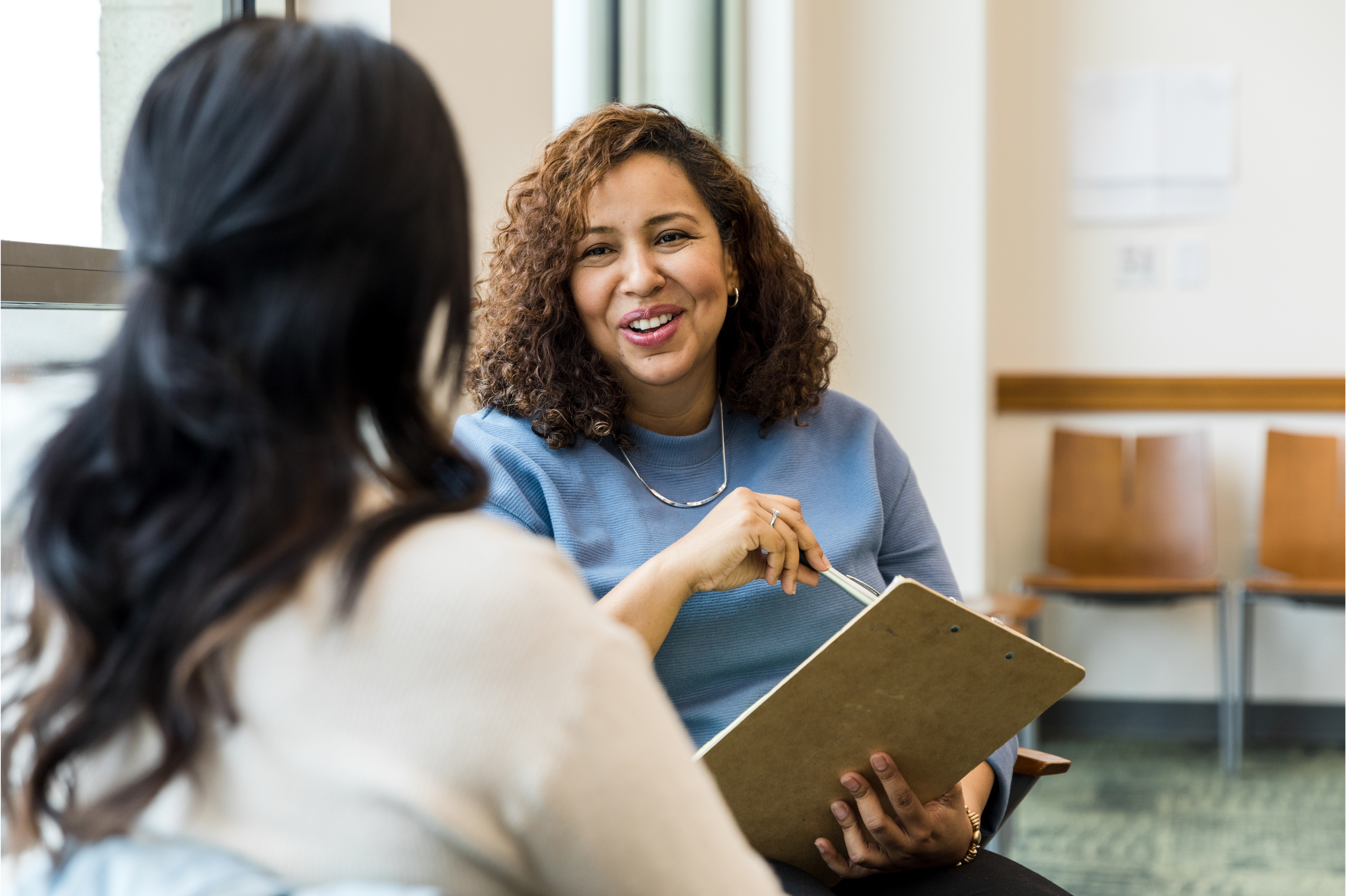 Photo of two people in an office setting. One person with long black hair has their back to the camera and is facing a woman with medium skin tone and curly brown hair, smiling as she holds a pen and clipboard.