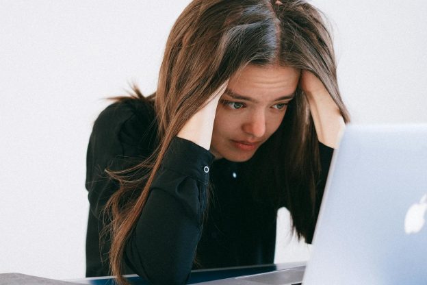 Photo of a light skinned woman straight brown hair, holding her head in her hands with a disressed look on her face. She is seated at a white table in front of an open Macbook.