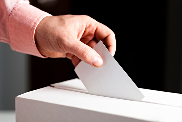 Photo of a person's hand with medium skin-ton and a pink sleeve visible as they deposit a ballot into the slot on the top of a white cardboard box.